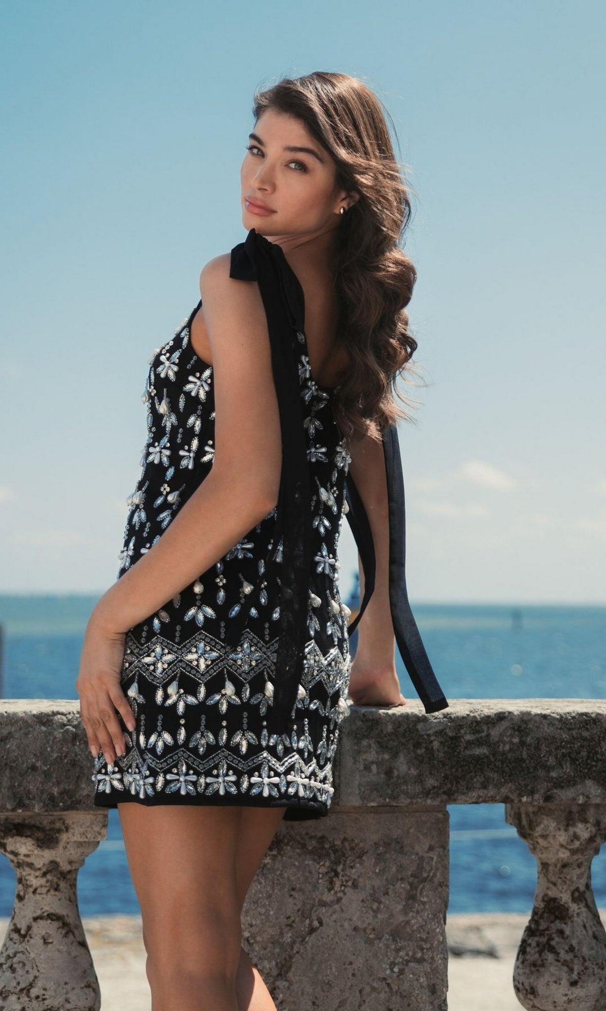 A woman in the Short Wedding Guest Dress 10937 by Mac Duggal stands by a stone railing, gazing over her shoulder with the ocean and sky in the background.
